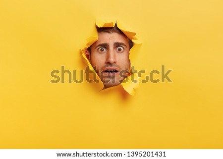 Human facial expressions concept. Worried young man with dark eyes, bristle, frowns eyebrows, has scared look, gazes through paper hole, feels nervous and puzzled. Yellow background with blank space
