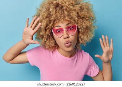 Human Facial Expressions And Reactions Concept. Curly Haired Woman Keeps Lips Folded Palms Raised Looks Surprisingly At Camera Wears Trendy Pink Sunglasses And T Shirt Isolated Over Blue Background