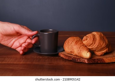 Human Female Hand Holding Black  Cup Of Espresso Coffee, Two Croissants, Rolls On Craft Pad On Wooden Background At Dark Blue Wall. Bakery Food, French Breakfast, Morning Menu, Cafe Concept. Side View