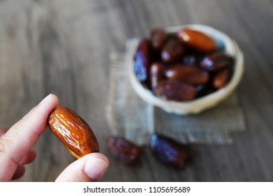 Human Finger Holding Single Date Fruit And Blurred Dates Filled Bowl In Background          