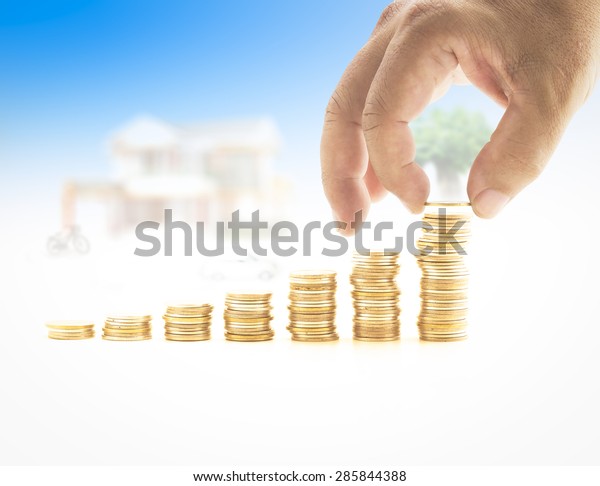 Human hand\
adding a golden coin in the final row of golden coins over blurred\
house and car on blue sky\
background