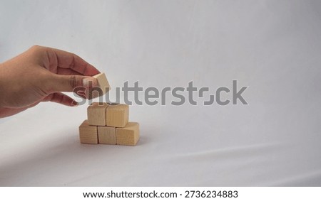 Human hand carefully stacking small wooden cubes, forming a stable structure on a plain white backdrop, Foundation Concept.