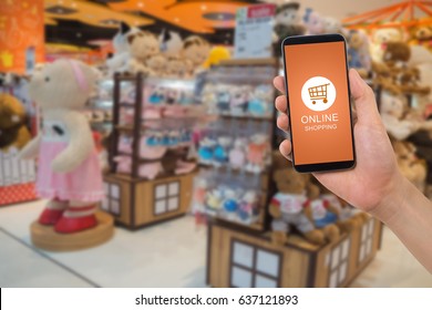 Human Hand Hold Smartphone, Tablet, Cell Phone With Online Shopping Application Screen On Blurry Toy Shop Background.