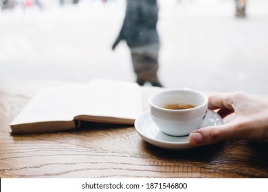 Human Hand Holding Black Coffee On Saucer With Vintage Book On Background On The Wooden Table Next To Big Coffee Shop Window And Looking To Passerby Street Walkers