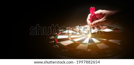 Human hand holding a darts hitting at the target on black background. Dart in the bullseye. Goal-oriented concepts, planning, and marketing strategies.