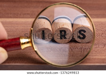 Human Hand Holding Magnifying Glass Over Wooden Cork With IRS Text On Wooden Textured