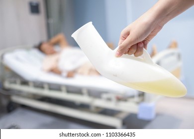 Human Hand Holding A Portable Urinal Plastic Over Blurred Patient On The Bed.