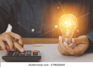 Human Hand Holds A Light Bulb And Presses A Calculator To Calculate The Company Energy Use Numbers. Saving Energy And Accounting Finance Concept.