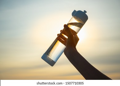 Human hand holds a water bottle against the setting sun. Close up of a reusable water bottle in a human hand, concept of thirst, rehydration and decreasing single use plastic