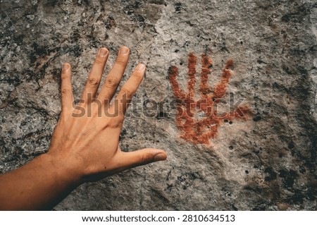 A human hand placed next to a red prehistoric handprint on a cave rock wall.