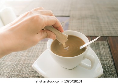 Human Hand Pouring Sugar To Cup Of  Hot Coffee, Focus On Packet Of Sugar