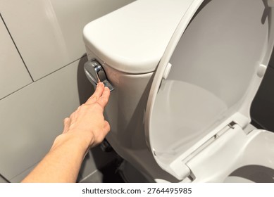 Human hand push chrome flush button on white toilet tank. Toilet lid is open. Clean modern bathroom with light gray tiled wall visible in background.