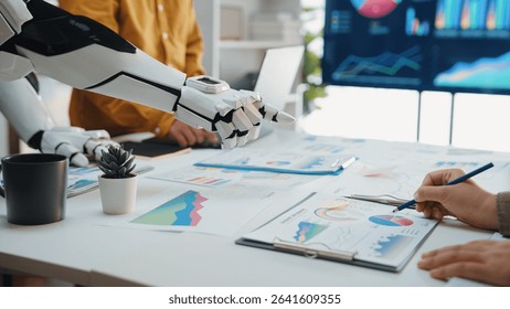 A human hand and a robotic arm collaborate on data analysis in a modern office space, showcasing innovative technology and teamwork through digital charts and graphs.Noogenesis - Powered by Shutterstock - Get 15% off with code: PIKWIZARD15