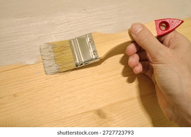 A human hand is using a paint brush to apply light gray or white paint to a raw wooden surface indoors, showing a clear contrast between the unfinished and finished wood planks