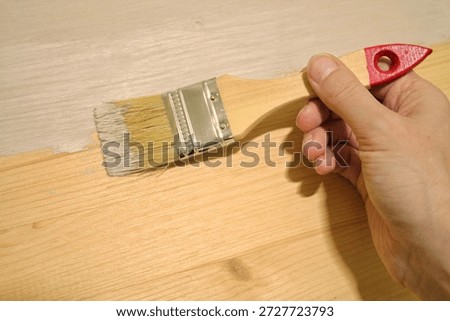 A human hand is using a paint brush to apply light gray or white paint to a raw wooden surface indoors, showing a clear contrast between the unfinished and finished wood planks