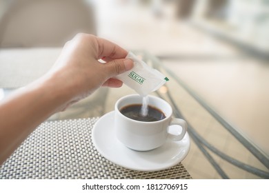 Human Hand With White Sugar Packet And Pouring In Hot Black Coffee , In Soft Focus