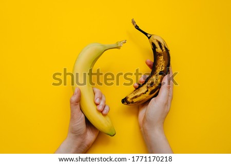 Human hands holding two bananas: fresh and ripe, on bright yellow background.