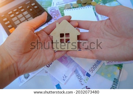 in human hands lies a small wooden house, against the background of dollar and euro bills.Calculator and coins. Concept - buying, selling real estate. Top view, horizontal photo.