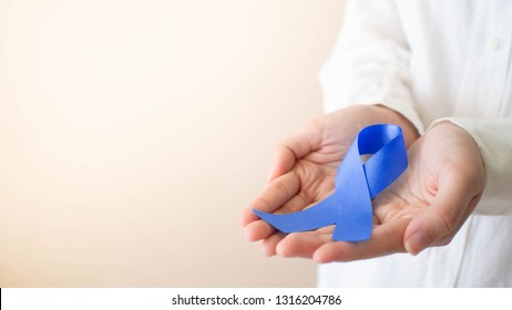 Human Hands In White Shirt Holding Dark Blue Ribbon Awareness Symbol For Support People Living W/ Colon Or Colorectal Cancer. Health Care And Medical Concept. Copy Space.