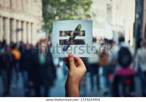 Human inequality as global social issue. Stop\
discrimination on grounds of race, sex or religion as hand holding\
a paper sheet with injustice, unfairness symbol over crowded street\
background.
