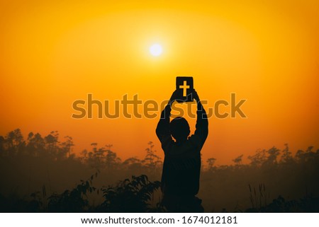 Human kneel down and lift the christian Bible up on head with light sunset background, christian silhouette concept.