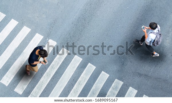 Human life\
in Social distance. Aerial top view with blur man with smartphone\
walking converse of other people at pedestrian crosswalk on grey\
pavement street road with empty\
space.