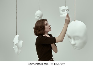 Human roles. A girl in black clothes stands in a white room among different masks deciding which one to choose. Hypocrisy. Mental disorders. - Shutterstock ID 2145567919