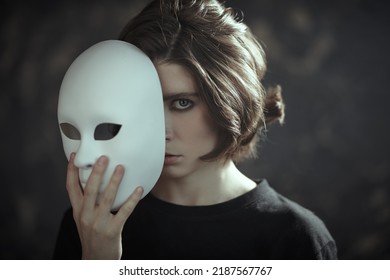 Human roles. Hypocrisy. Portrait of a brunette girl in black clothes peeking out from behind a mask-role in her hand on a dark grunge background. Mental disorders. 
