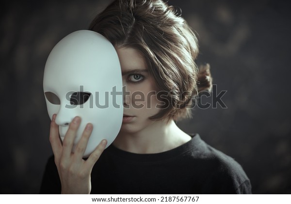 Human roles. Hypocrisy.\
Portrait of a brunette girl in black clothes peeking out from\
behind a mask-role in her hand on a dark grunge background. Mental\
disorders. 