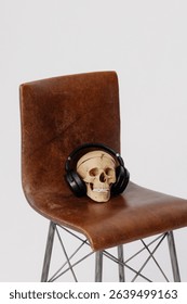Human skull with black headphones placed on vintage brown leather chair with metal legs, white wall background. Concepts: surrealism, music, mortality, creative contrast.