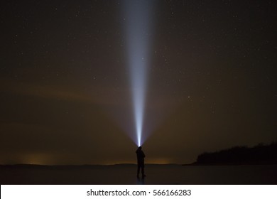 Human Standing On Ice Lake At Night In Swedish Winter Landscape.Shining Up In Sky With Flashlight At Stars.Beautiful Blue Light Beam. Nice, Calm And Peaceful Image. Lovely Abstract Photo.