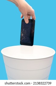 Human Throws A Broken Mobile Phone Into A Trash Can On A Blue Background. Human Hand Holds A Phone With A Broken Screen Over A White Trash Can And Prepares To Throw Away The Broken Equipment