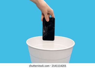 Human Throws A Broken Mobile Phone Into A Trash Can On A Blue Background. A Human Hand Holds A Phone With A Broken Screen Over A White Trash Can And Prepares To Throw Away The Broken Equipment