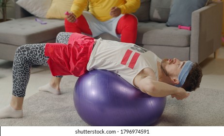 Humor In Sports. Fat African American Lazy Guy Resting On Couch While His Clumsy Funny Friend Doing Fake Gymnastics With Fitness Ball.