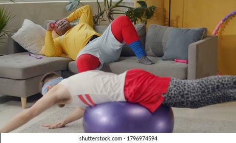 Humor In Sports. Fat African American Lazy Guy Resting On Couch While His Clumsy Funny Friend Doing Fake Gymnastics With Fitness Ball.