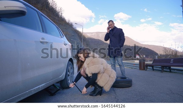 humor. woman changing a car wheel. man talking on\
the phone.