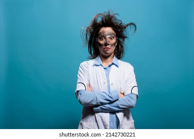 Humorous mad scientist with erratic hair and filthy face standing with crossed arms. Hilarious looking crazy chemist with messy hairstyle and dirty face standing on blue background looking at camera.