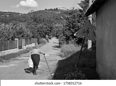 Hunched old woman walking with a stick down the country road. Elderly woman with a cane next to school traffic sign in a village. Black and white image, diseases of the spine and concept of old age. 