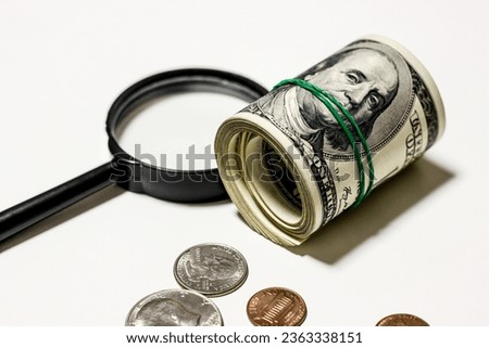 Hundred dollar bills are rolled up with rubber band with coins and magnifying glass on white background