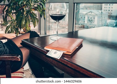 A Hundred Dollars Generous Tip On The Table In A Leather Folder. The Girl Drank The Wine And Paid The Bill. A Large Wine Glass On The Table In An Expensive Restaurant.