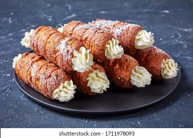Hungarian dessert kurtos kalacs or chimney cake filled with cream sprinkled with icing sugar on top served on a black plate on a dark concrete background, horizontal orientation, close-up