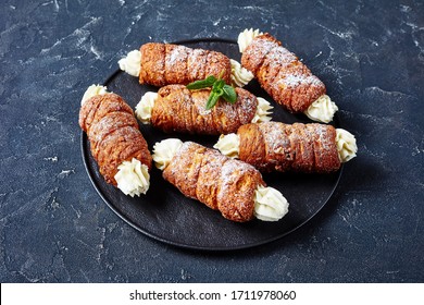 Hungarian kurtos kalacs or chimney cake filled with buttercream decorated with fresh mint on top served on a black serving tray on a dark concrete background, close-up