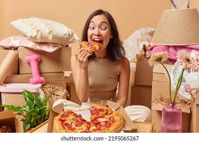 Hungry adult Asian woman bites slice of tasty pizza has funny expression dressed casually poses on floor around cardboard boxes full of personal belongings prepares for moving and relocation