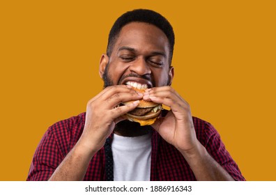 Hungry African American Guy Eating Burger Standing On Yellow Background In Studio. Black Man Enjoying Junk Food. Binge Eating And Unhealthy Nutrition, Fast Food Overeating Concept