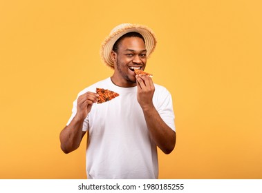 Hungry African American Guy In Trendy Summer Outfit Eating Pizza, Having Snack On Yellow Studio Background. Positive Black Man In T-shirt And Straw Hat Enjoying Fast Food