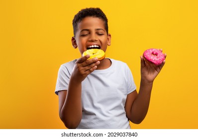 Hungry African American Teen Boy Holding Colorful Donuts, Eating Junk Unhealthy Food Full Of Sugar, Isolated On Yellow Studio Background, Copy Space. Kids Obesity And Overweight Concept