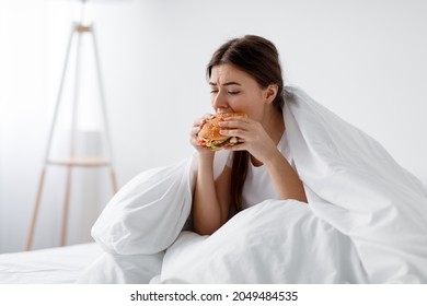 Hungry After Diet, Starving Stressed Caucasian Millennial Woman Eating Burger In Bed At Home Alone. Unhealthy Overeating Lifestyle, Depression, Psychological And Mental Problems. Eating Too Much