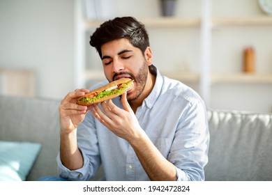 Hungry Arab Man Eating Sandwich With Eyes Closed, Enjoying Delicious Fast Food At Home