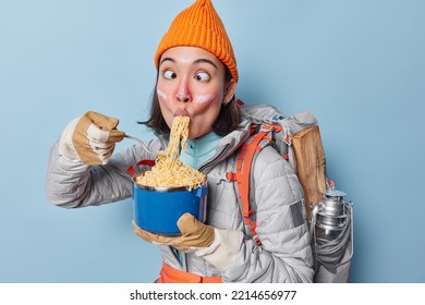 Hungry Asian Female Tourist Eats Noodles Rests After Hiking Trip Or Long Journey Carries Backpack With Equipment Makes Funny Grimace Crosses Eyes Wears Orange Hat Jacket And Gloves Poses Indoor