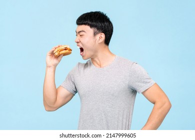 Hungry Asian Man In Casual Wear Eating Fresh Unhealthy Burger While Standing On Light Blue Isolated Background In Studio